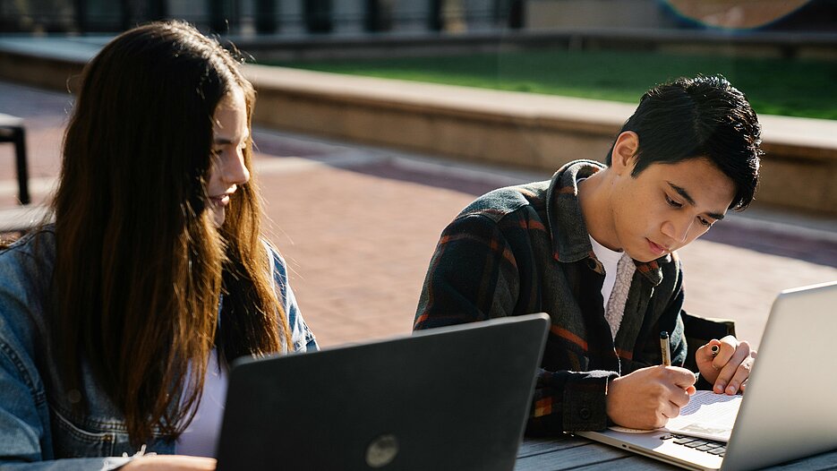 Zu sehen sind zwei junge Menschen, die draußen an Laptops arbeiten