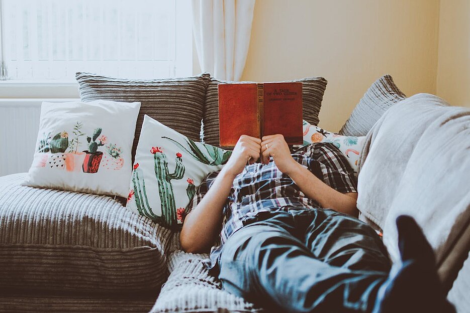 Eine Person liegt lesend auf der Couch. Die Füße zeigen in Richtung Kamera, das Gesicht ist vom Buch verdeckt.