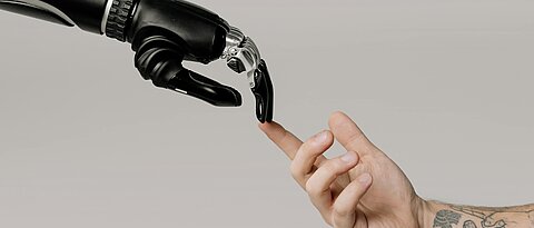 eine Roboterhand und eine menschliche Hand stellen die Hände von Gott und Adam aus dem Gemälde "die Erschaffung Adams" von Michelangelo nach