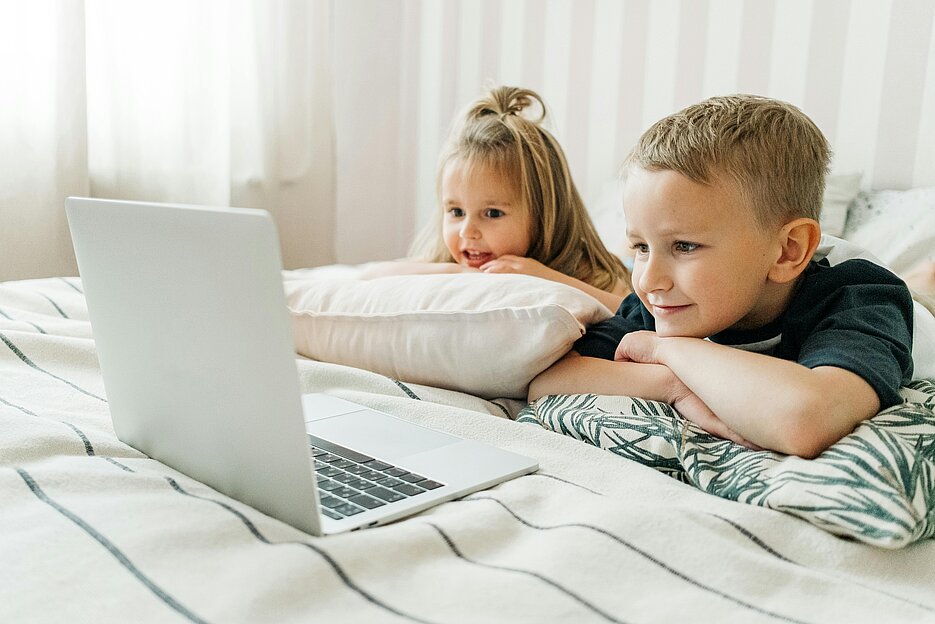 Zwei Kinder liegen auf dem Bett und schauen auf einen Laptopbildschirm